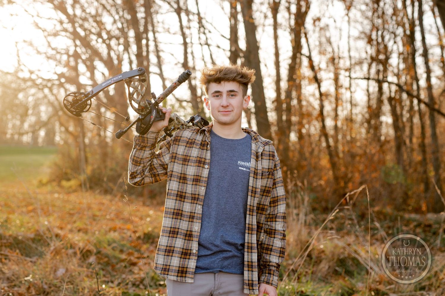 Person holding compound bow stands in wooded area during sunset wearing plaid shirt.