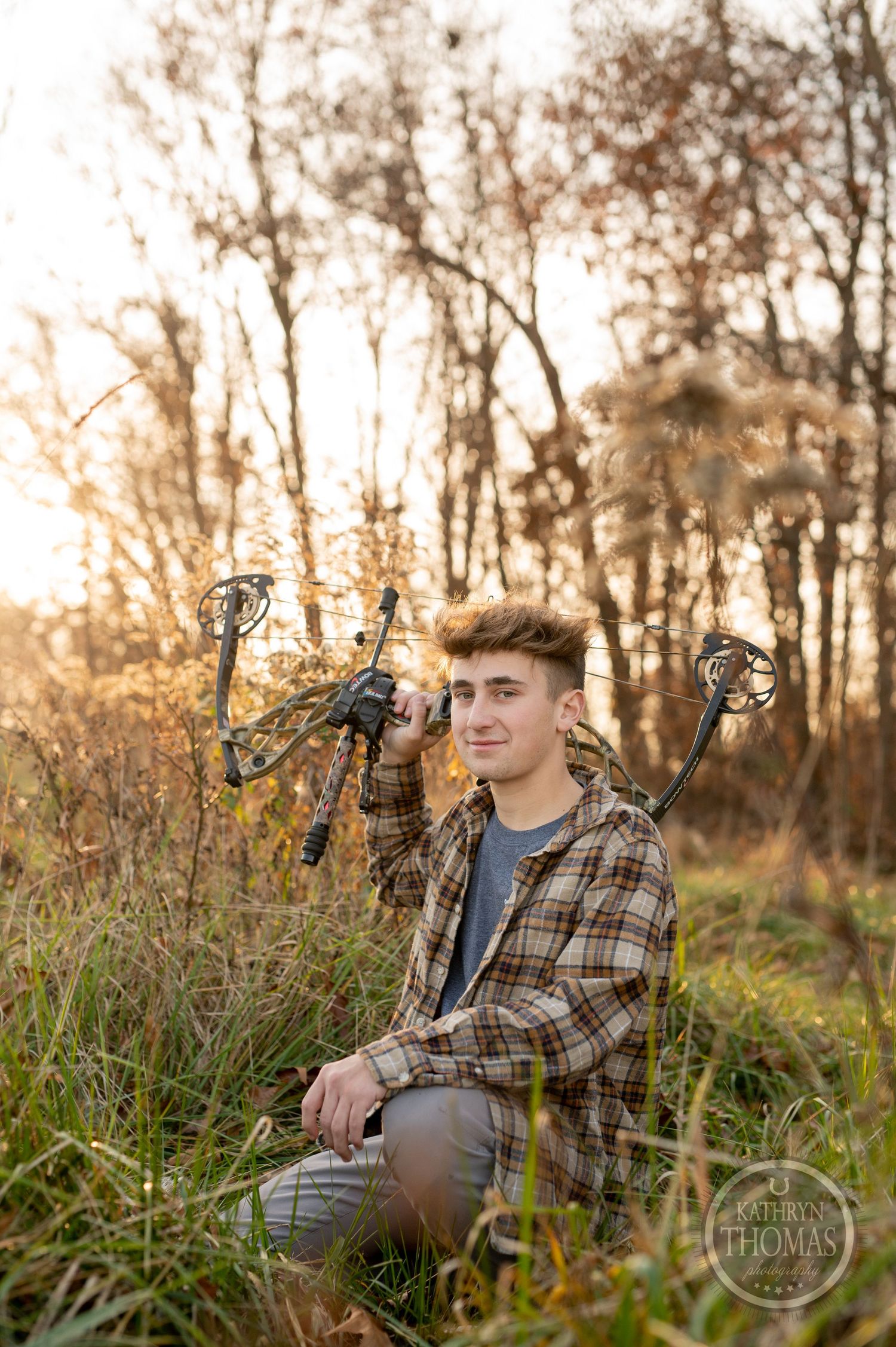 Archer poses with compound bow while kneeling in autumn meadow at sunset.