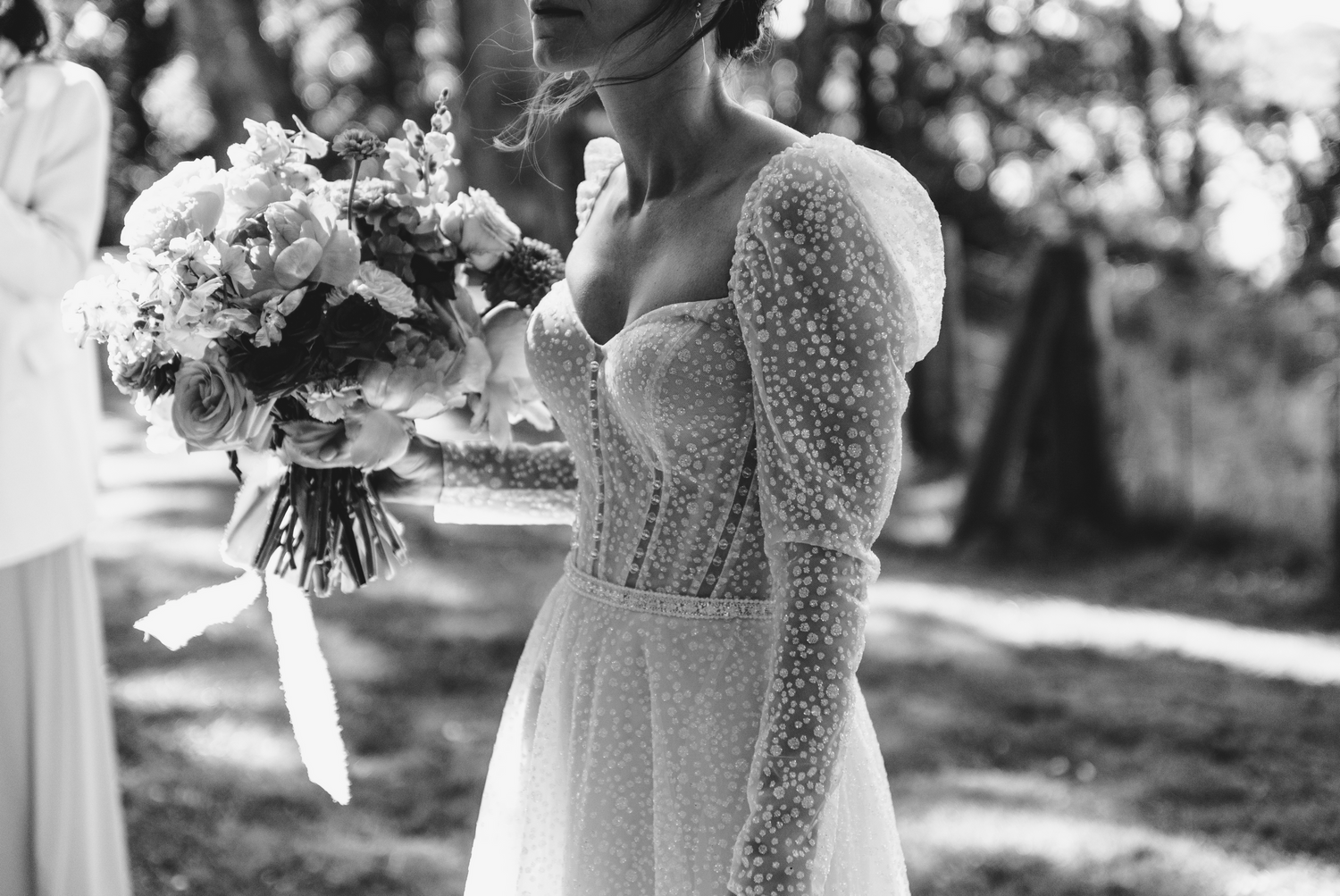 Une photographie en noir et blanc montre une robe de mariée en dentelle détaillée avec des manches longues et un bouquet de fleurs.