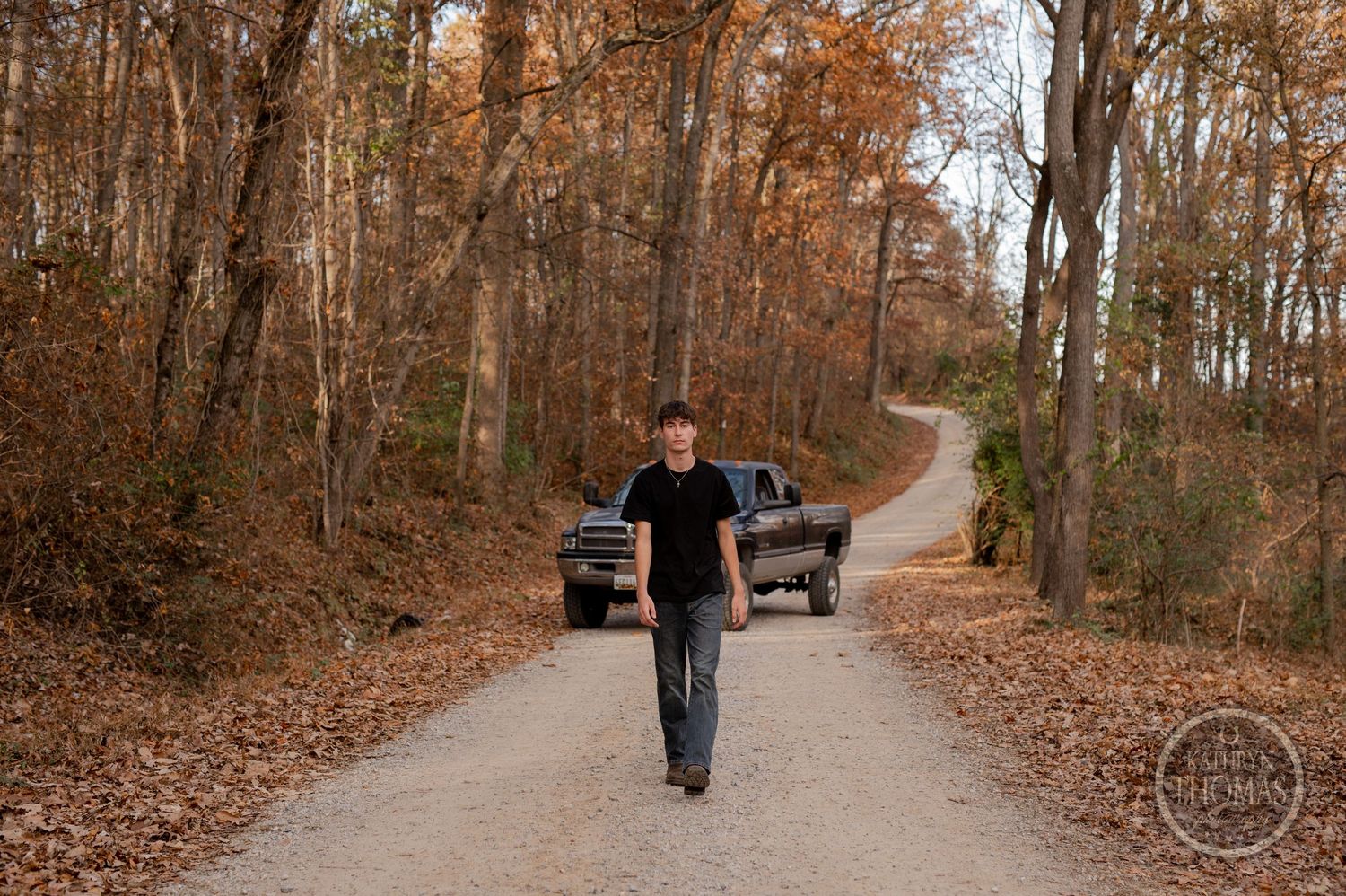 Person walking down winding dirt path with truck visible in autumn woods.