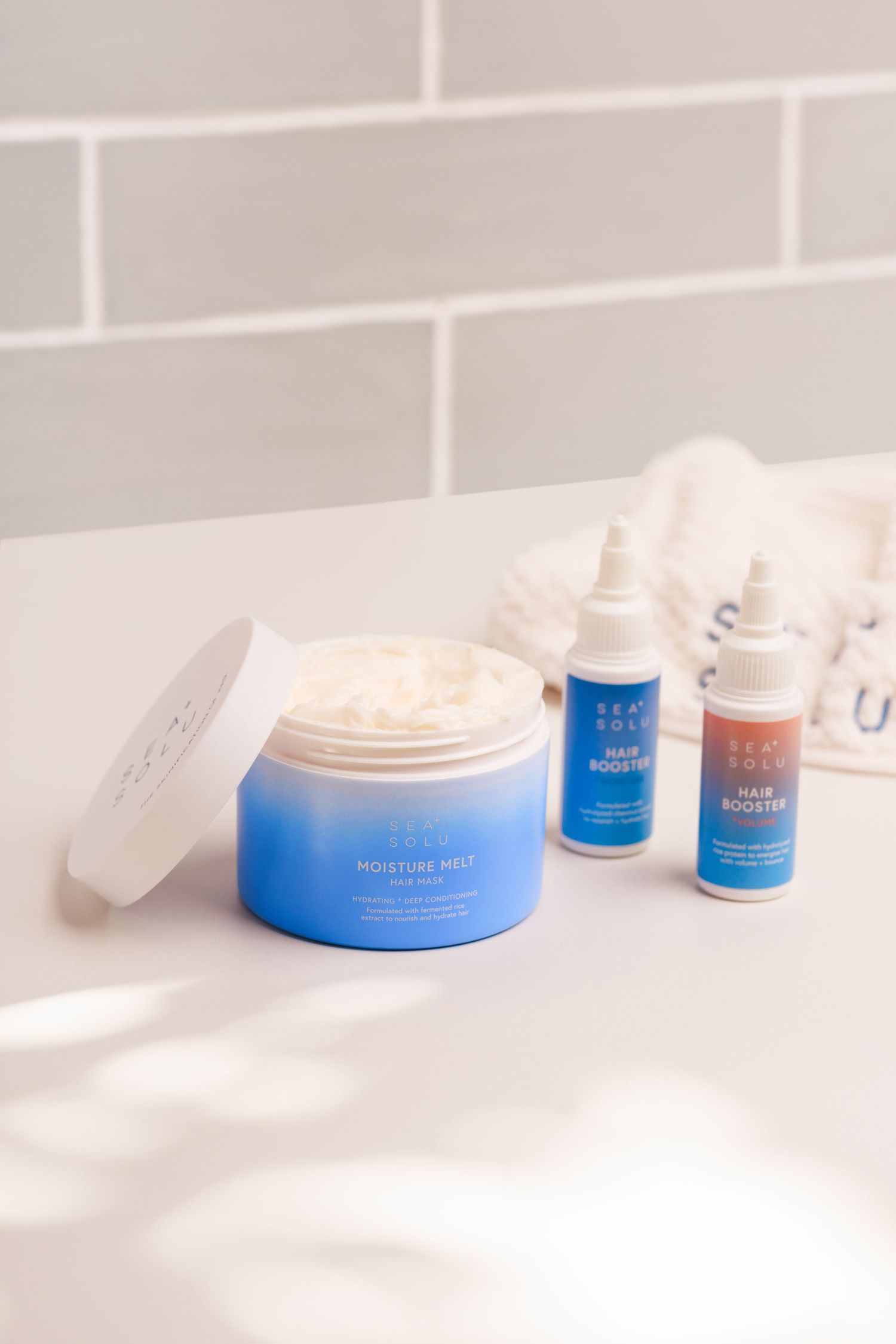 Blue hair mask jar and booster serum bottles displayed on white bathroom counter with a brand hair towel. There is leafy light and shadows.