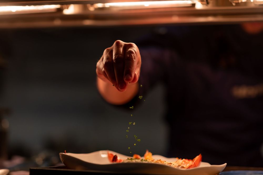 Close-up of someone seasoning a dish with spices falling from their hand in a dark restaurant setting.