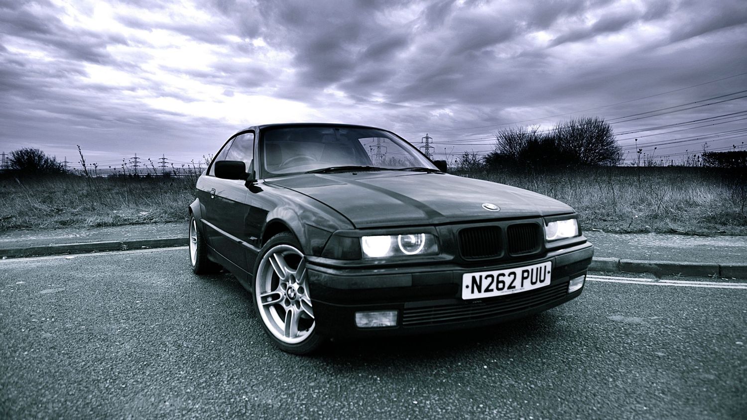 Black BMW E36 coupe parked on a road under dramatic cloudy skies.