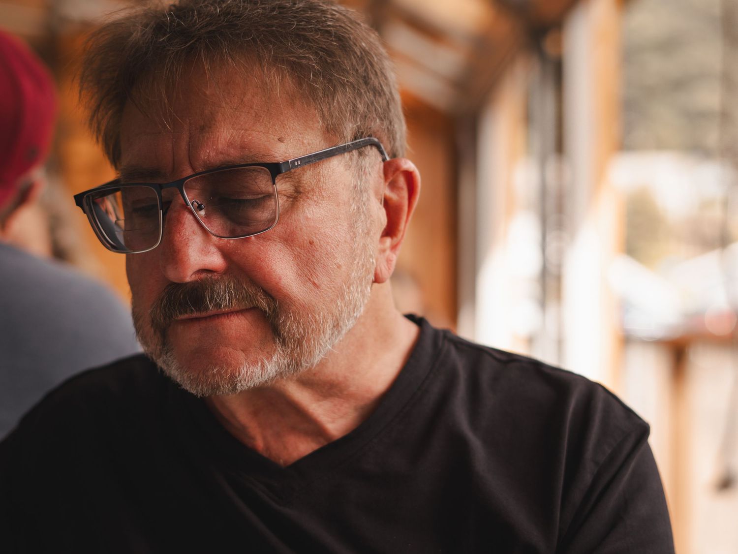 A contemplative portrait shot showing a person wearing glasses in a black shirt against a warm-toned background.
