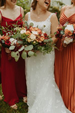 Bridesmaids in red and rust colored dresses hold lush floral bouquets next to bride in white gown.