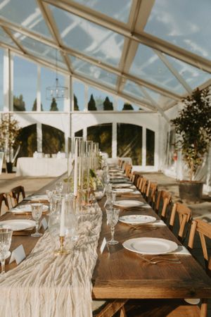 Elegant wedding reception table setup under clear tent with rustic wooden chairs and white place settings.