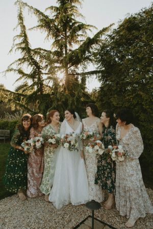 A bridal party in floral dresses poses together outdoors at sunset under tall trees.