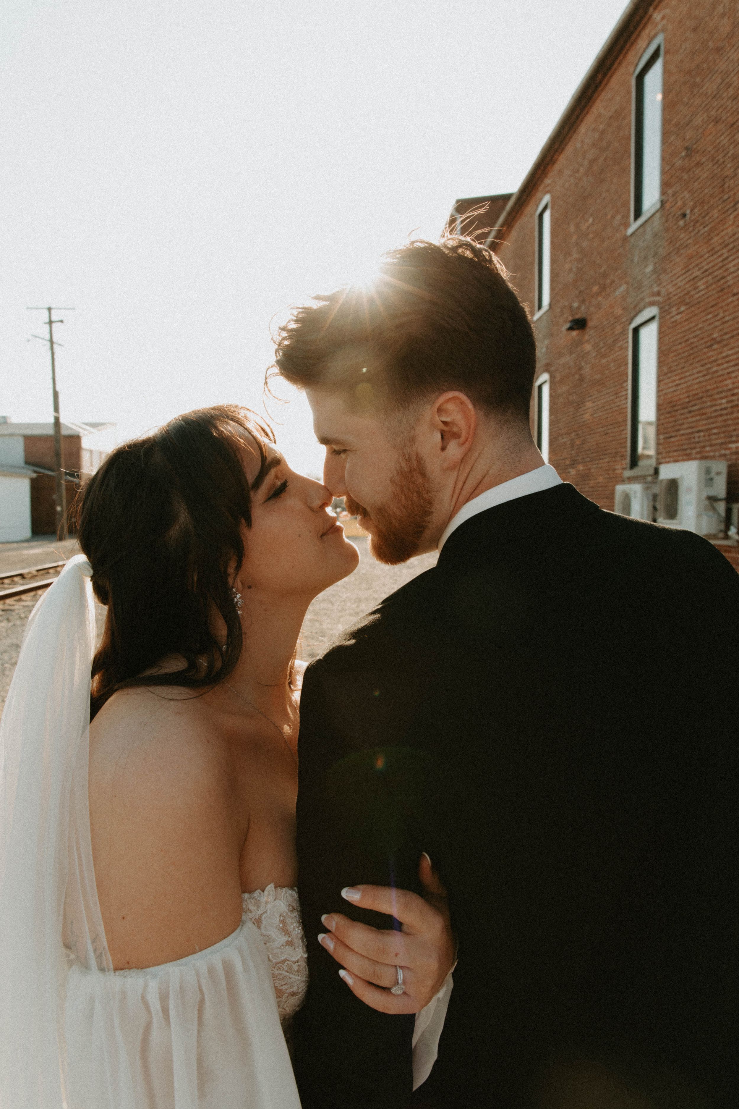 A romantic sunset moment between newlyweds sharing a kiss against a brick building.