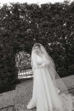 A bride in a flowing white dress and veil poses elegantly against a hedge backdrop.