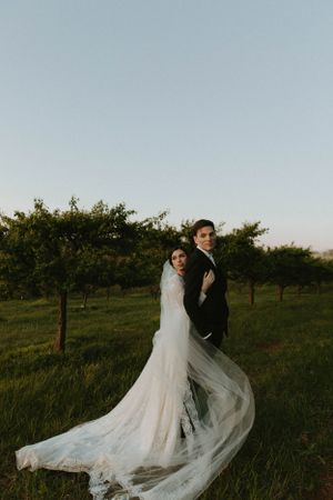 Bride's dress train flows dramatically during outdoor orchard photoshoot.