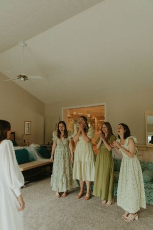Bridesmaids in vintage-style dresses react with joy and excitement in a bedroom setting.