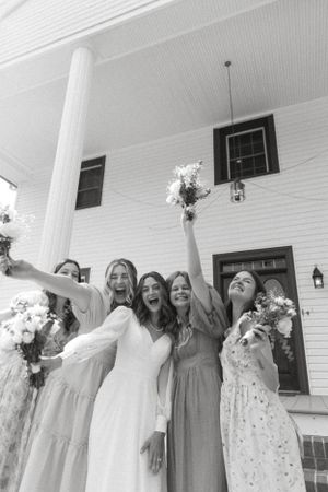 Black and white photo of bridal party celebrating on a white porch with raised bouquets.
