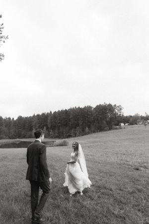 Black and white wedding photo of couple walking through a scenic field with trees.