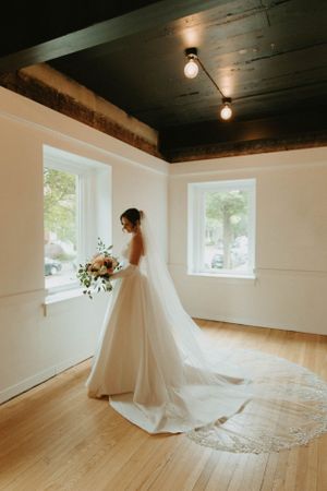 A bride in a flowing white gown with long train stands by windows in a sunlit room.