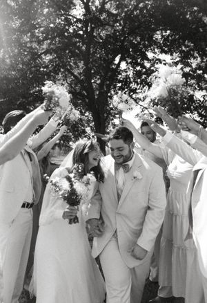 Wedding couple celebrates with guests raising arms under a tree during outdoor ceremony in black and white.