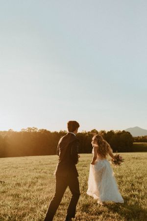 A couple runs through a sunlit field at golden hour in wedding attire.