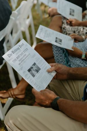 Wedding guests holding ceremony programs while seated in white folding chairs.