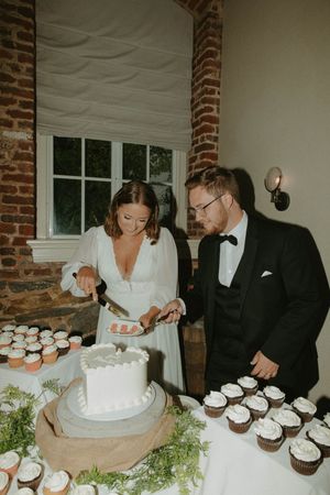 Newlyweds cutting their wedding cake surrounded by cupcakes at rustic brick venue.