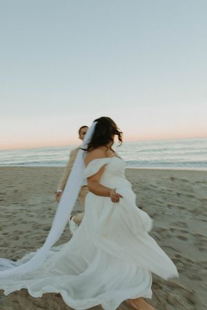 Bride in flowing white dress twirls on beach at sunset with ocean waves in background.