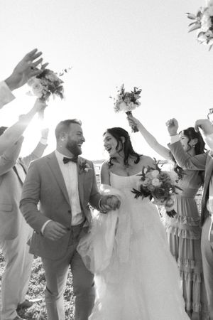 Joyful wedding celebration with guests tossing bouquets in the air while couple embraces under bright sunlight.