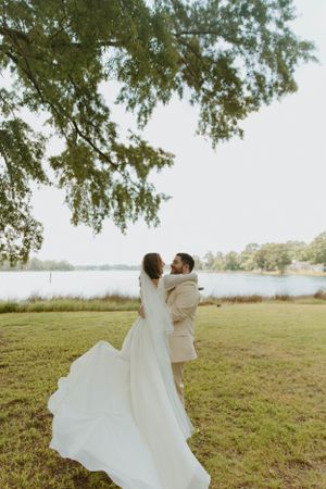 Bride in flowing white gown embraces groom by lakeside under overhanging trees.