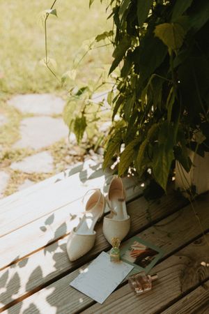 White bridal shoes and stationery arranged on wooden deck with dappled sunlight.