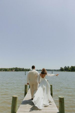 Newlyweds in elegant attire walk together on a wooden dock overlooking a serene lake on their wedding day.