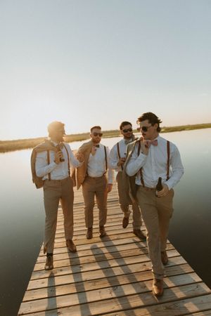 Groomsmen in suspenders sharing laughs while standing on a wooden dock at sunset.