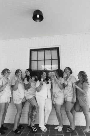 Wedding party in matching outfits poses playfully against a white brick wall in black and white.