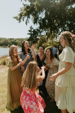 Joyful wedding guests laugh and celebrate together outdoors by a lake on a sunny day.
