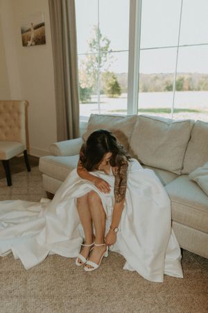 A bride in a white gown sits on a couch while putting on her wedding shoes.