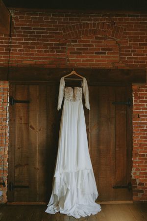 Elegant white wedding dress hanging against rustic wooden doors and brick wall.