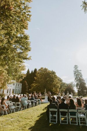 An outdoor wedding ceremony setup with white chairs on a lawn under tall trees.