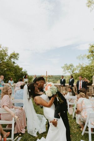 A romantic outdoor wedding ceremony with guests sitting on white chairs as the couple shares their first kiss.