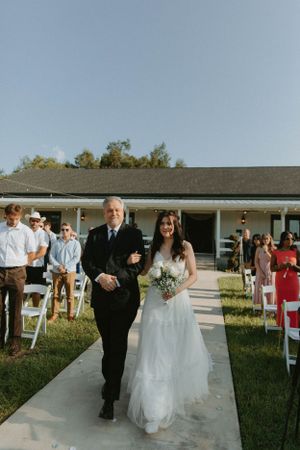 A traditional wedding processional down an outdoor aisle with white chairs and guests.