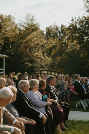 Wedding guests seated in rows of white chairs during an outdoor ceremony at sunset.