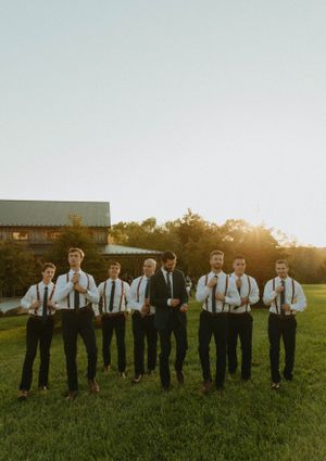 A group of groomsmen in matching suits and suspenders pose together at sunset on a grassy field.