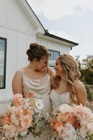 Two people sharing a joyful moment with peach and blush floral bouquets against white building.