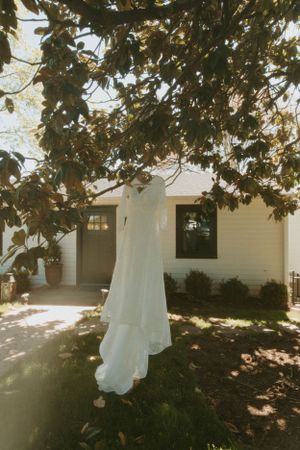 A white wedding dress hangs from a tree branch outside a cream-colored house with classic architecture.