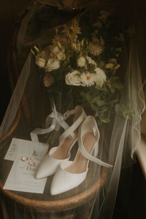 Wedding accessories including white heels, veil and floral bouquet arranged on wooden surface.