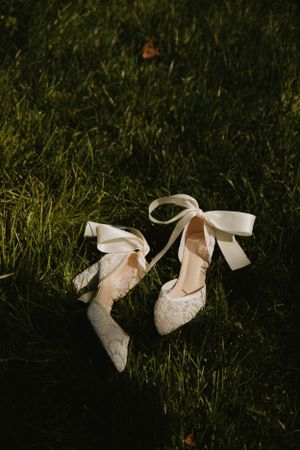 Elegant lace wedding shoes with ribbon ties laying on grass in natural lighting.