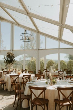 Light-filled reception venue with exposed beams, chandeliers and cross-back chairs at round tables.