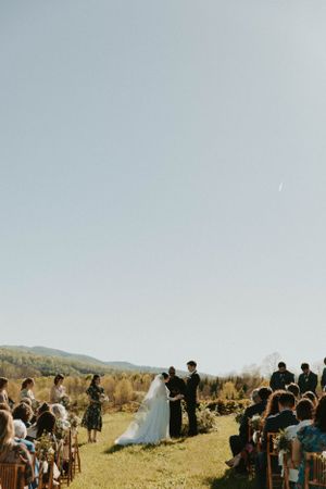 Outdoor wedding ceremony in a mountain landscape with guests seated on wooden chairs.