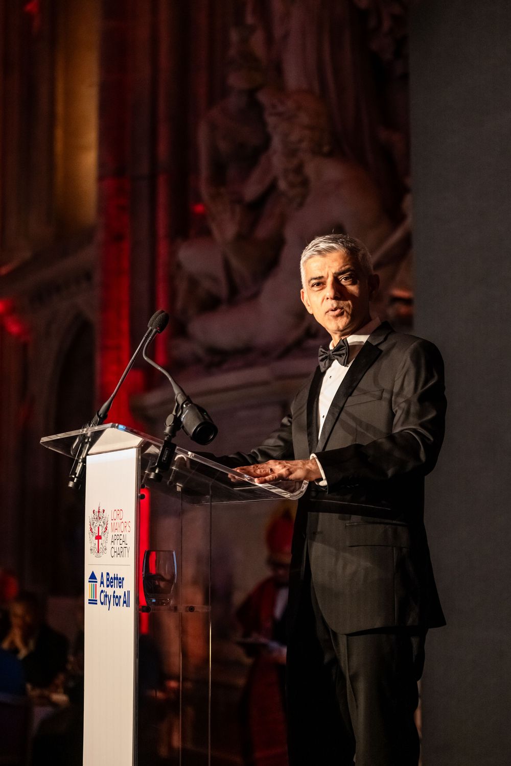 A man in a dark suit stands at a podium giving a speech in a dimly lit venue with red lighting.