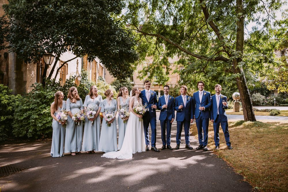 Wedding party in blue suits and light gray dresses pose together on a tree-lined pathway.