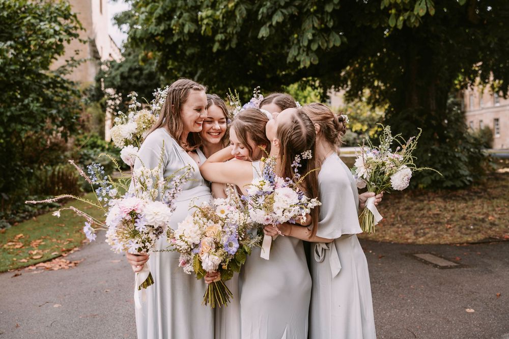 Bridesmaids in pale gray dresses share a joyful moment while holding pastel bouquets.