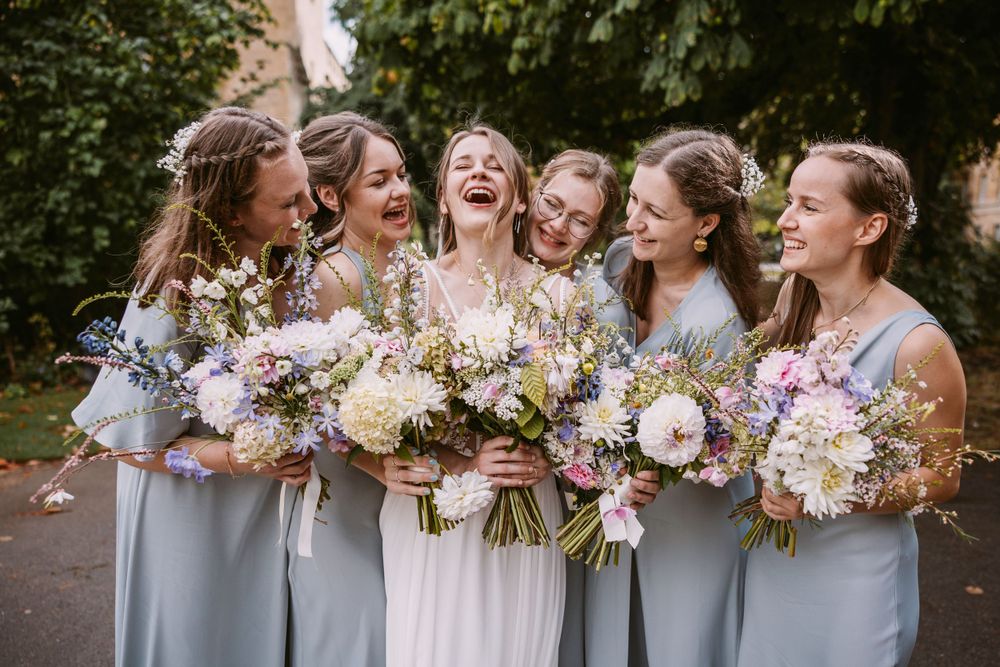 Wedding party members share genuine laughter while holding romantic pastel flower bouquets.