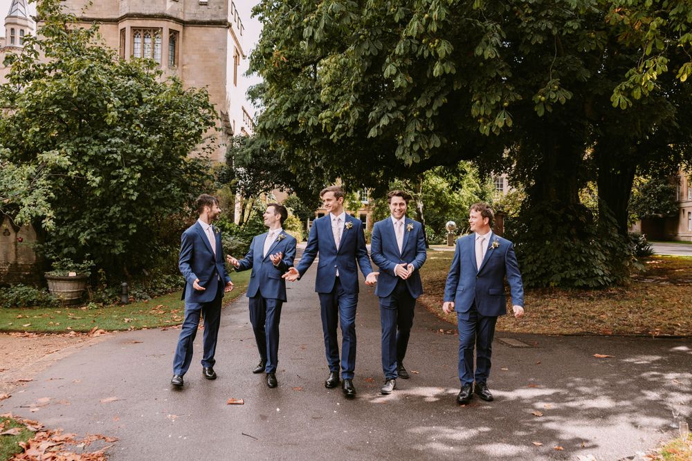 Groomsmen in navy suits walk together on garden path near historic building at Balliol College, Oxford