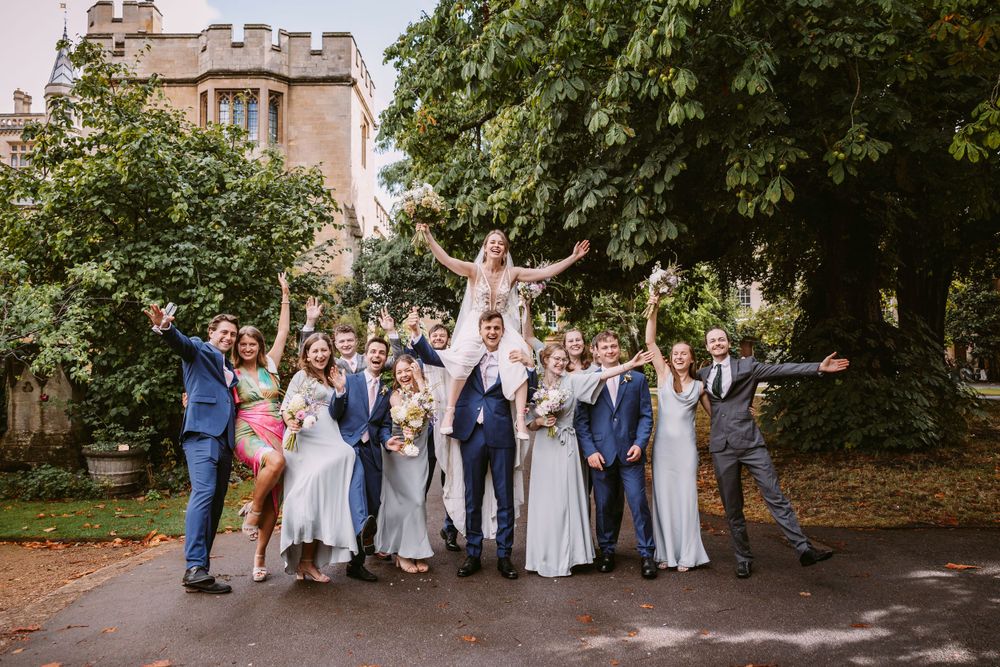Wedding party celebrates with arms raised on path near historic stone building at Balliol College, Oxford