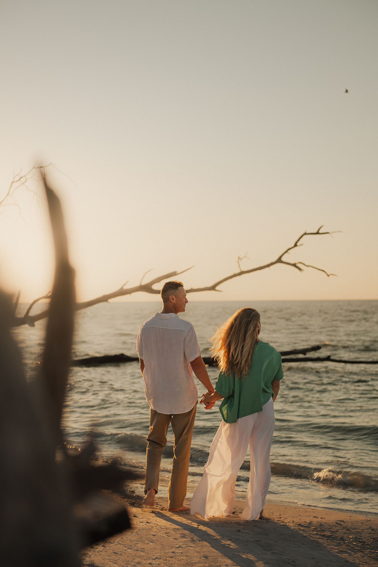 Couple stands on beach at sunset watching ocean waves with driftwood branch framing scenic coastal view.